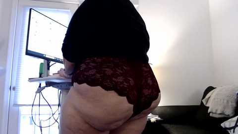 Media: Video of a plus-sized woman in maroon lace panties, standing in a dimly lit room with a computer desk and a white wall.