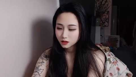 Media: Video of an East Asian woman with long black hair and fair skin, wearing red lipstick, sitting indoors. She has tattoos on her right shoulder and arm. Background includes a floral-patterned bedspread and a dark room with dim lighting.