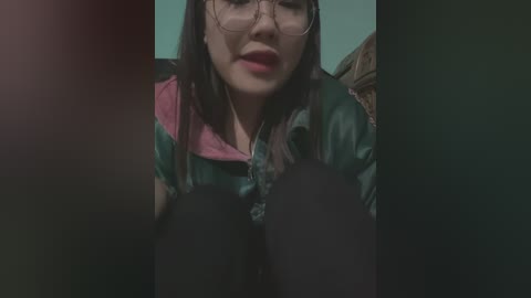 Media: Video of a woman with light skin, long dark hair, and large glasses, wearing a colorful shirt and black leggings, sitting in a dimly lit room.