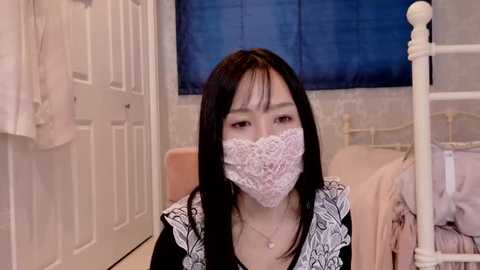 Media: Video of an Asian woman with long black hair, wearing a white lace mask, black dress, and white apron, sitting in a bedroom with a white bed and blue window blinds.