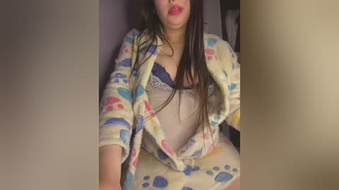 Media: Video of a woman with long brown hair, wearing a pink and blue paw print bathrobe, exposing a blue lace bra underneath. She has light skin and is sitting on a bed with beige walls in the background.