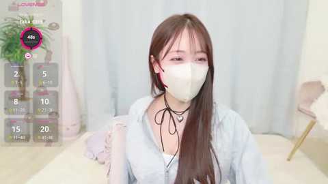 Media: A video of a young Asian woman with long brown hair, wearing a white mask, light blue hoodie, and black choker, standing in a soft, pastel-colored room.