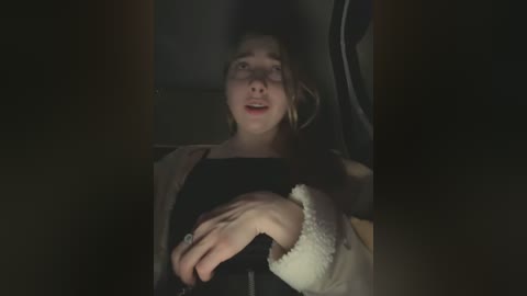 Media: Video of a young woman with light skin, brown hair, and a light jacket, lying in a dark car, with a look of surprise on her face.