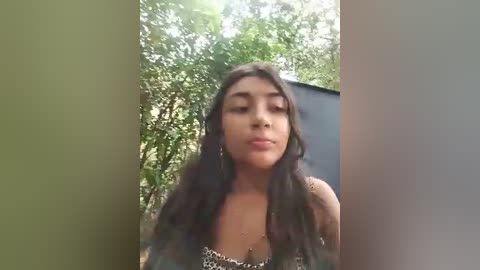 Media: A video of a young woman with long, wavy black hair, wearing a leopard-print top, standing in a lush, green outdoor setting with trees and a blue wall in the background.