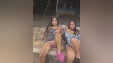 Media: Video of two young women with long black hair, sitting on a bench, wearing tight, revealing outfits, one in pink, the other in denim. Background shows a dark, urban setting.