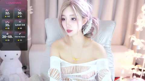 Media: Video of a fair-skinned, slender Asian woman with long, platinum blonde hair, wearing a white, off-shoulder mesh top, sitting on a light-colored couch. Background features a teddy bear and soft, pastel curtains.