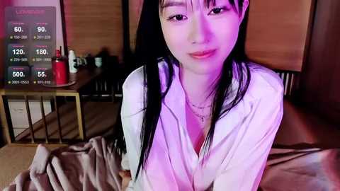 Media: Video of an East Asian woman with long black hair, wearing a white shirt, lying on a bed with a TV showing a \"Live\" stream. Background includes a wooden wall and a nightstand with a red bottle.