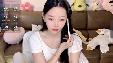 Media: Video of an Asian woman with long black hair, wearing a white t-shirt, sitting on a brown couch surrounded by stuffed animals.