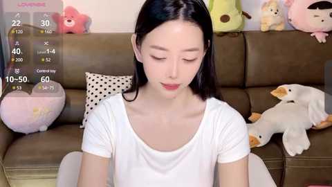 Media: Video of an East Asian woman with long black hair, fair skin, and closed eyes, sitting on a brown couch adorned with stuffed toys.