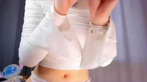 Media: Video of a woman in a white, long-sleeved crop top, revealing her toned midriff and navel, with a silver bracelet. Background shows a soft-focus, muted color palette of pastel shades.