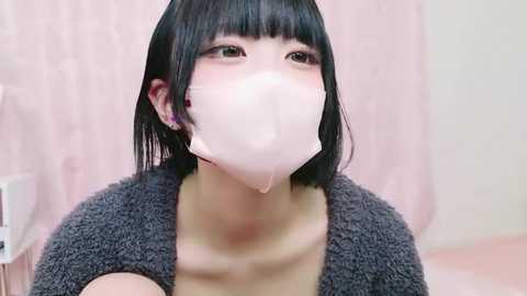 Media: Video of a young Asian woman with short black hair, wearing a gray knitted sweater and a pink surgical mask, looking upward, in a pale pink room.