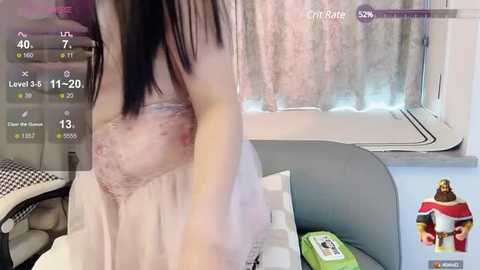 Media: A video of a woman with long black hair, wearing a sheer, pink lace dress, revealing her small, perky breasts, sitting in a modern living room with white curtains and a green cushion.