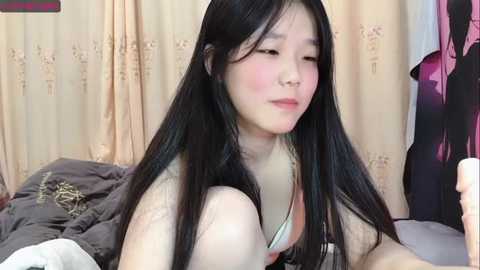 Media: Video of a young Asian woman with long black hair, wearing a white bra, lying on a messy bed in a dimly lit room.