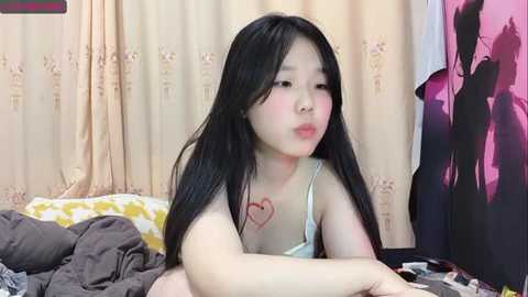 Media: A video of a young Asian woman with long black hair and fair skin, wearing a light-colored bra, lying on a bed with a heart tattoo on her chest. Background shows a beige curtain and a purple jacket hanging.