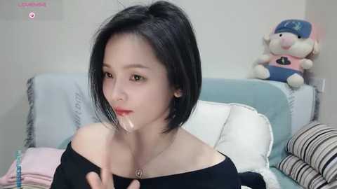 Media: Video of an Asian woman with short black hair and fair skin, wearing a black off-shoulder top, licking a phallic-shaped object, in a bedroom with a stuffed animal and pillows.