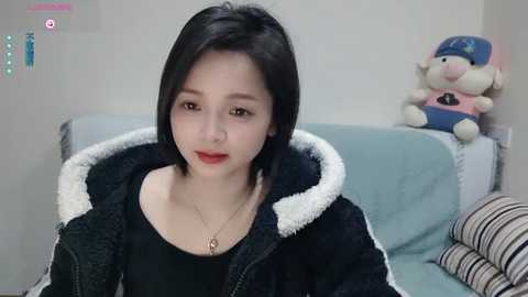 Media: Video of a young East Asian woman with short black hair, fair skin, and red lipstick, wearing a black hoodie with white fur lining, sitting on a light blue couch with a stuffed toy.