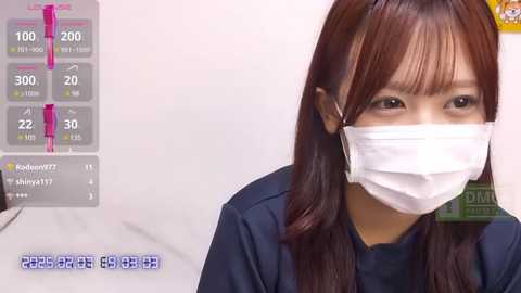 Media: Video of an Asian woman with long, brown hair wearing a white medical mask, navy blue scrubs, and a stethoscope, standing in a sterile, white hospital room with a monitor displaying vital signs and a heart rate of 122 bpm.