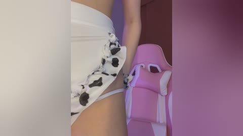 Media: Video of a close-up of a woman's midsection in white panties with black cow print, gripping a pink gaming chair, in a dimly lit room with purple lighting.