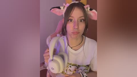 Media: Video of a young woman with light skin, wearing a white T-shirt, pink bunny headband, and white over-ear headphones, indoors with a pinkish hue.