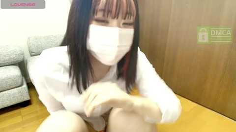 Media: Video of an Asian woman with long black hair and bangs, wearing a white face mask and long-sleeved shirt, sitting on a wooden floor in a living room with a gray couch and wooden wall in the background.