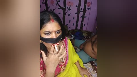 Media: Video of a South Asian woman in a yellow sari with red embroidery, wearing a black mask, smoking a cigarette, in a dimly lit room with floral-patterned purple wallpaper.