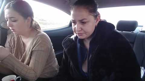 Media: Video of two women in a car: a fair-skinned woman with brown hair in a beige sweater and another with auburn hair in a dark coat, both looking down.