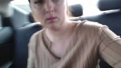 Media: Video of a woman with fair skin, brown hair, wearing a beige sweater, sitting in a car. Background shows car interior with blurred seats and windows.