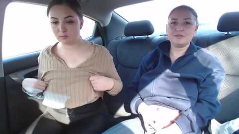 Media: Video of two women, one with a ponytail, in a car. Both are wearing casual jackets; one adjusts her seat belt, while the other sits calmly. The car's interior is visible, with beige and blue seats.