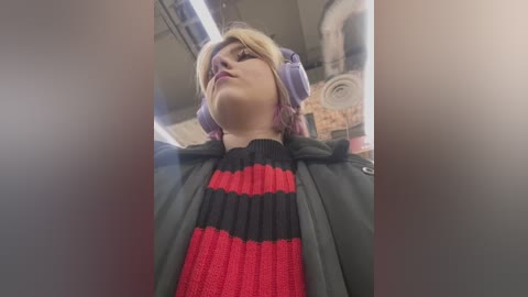 Media: A video of a young person in cosplay, with pale skin, blonde hair, and purple hair extensions. They wear large headphones and a red and black striped sweater, standing in a dimly lit, industrial room with exposed pipes.