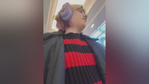 Media: A video of a young woman with light skin, pink hair, and large purple headphones. She wears a black and red striped sweater under a black jacket. Background shows a modern, indoor setting with bright lights.