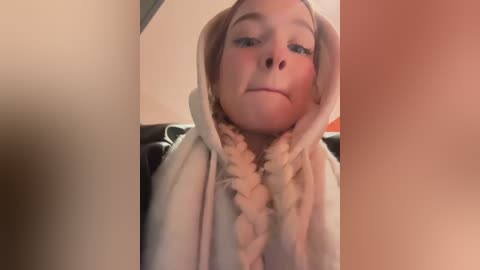 Media: Video of a young child with pale skin, dressed in a beige hooded sweatshirt with a fluffy, cream-colored braided scarf covering their face, leaving only their eyes visible.