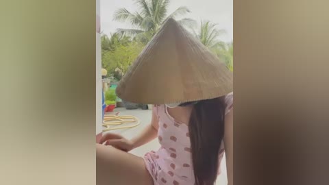 Media: Video of a woman wearing a conical hat, pink pajamas with heart pattern, seated outdoors with blurred background of palm trees and garden tools.