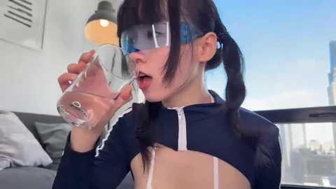 Media: Video of a young East Asian woman in a black sports bra, blue face mask, and glasses, drinking water in a modern, well-lit living room.