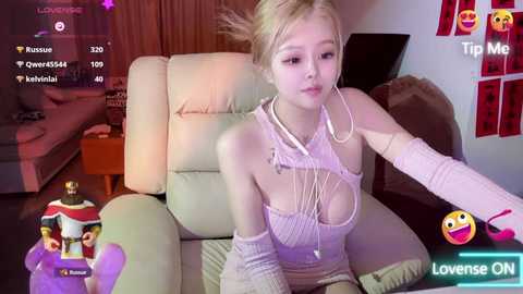 Media: Video of a young, fair-skinned, blonde woman with a slender physique and medium-sized breasts, wearing a revealing white crop top and long arm sleeves, sitting on a beige couch in a cozy living room.