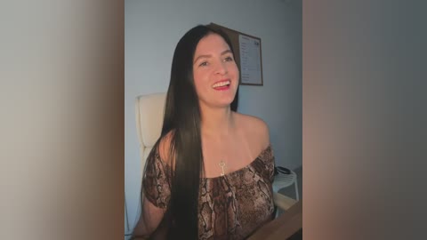 Media: A video of a smiling Asian woman with long black hair, wearing a brown snake-patterned off-shoulder top, seated in a white office chair. The background features a light blue wall with a framed document.