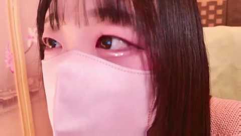 Media: Video of an Asian woman with straight black hair, wearing a white surgical mask, looking to the left, with a blurred background of a warm, indoor setting.