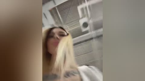 Media: Video of a blonde woman with a tattooed arm, partially obscured by a blurred foreground, standing in a white-tiled bathroom with visible plumbing and a window.