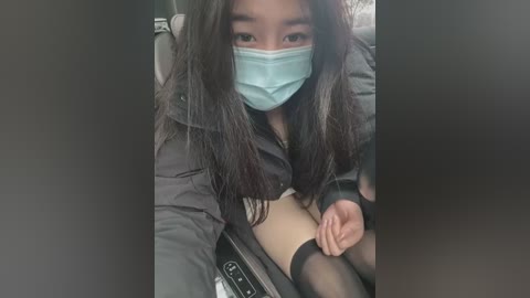 Media: Video of an East Asian woman with long black hair, wearing a blue face mask, black jacket, and sheer black thigh-high stockings, sitting in a car, partially visible wrist restraints on her right wrist.