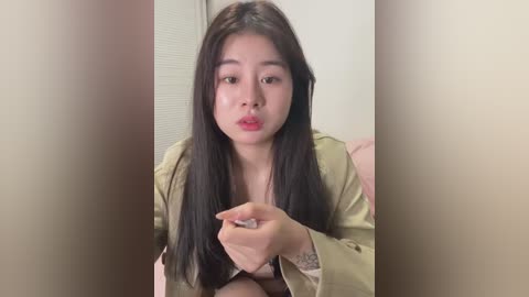 Media: A video of an East Asian woman with long, straight black hair and fair skin, wearing a beige jacket, sitting indoors with a pink background. She has a neutral expression and slightly parted lips.