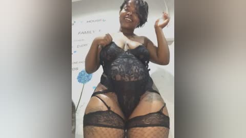 Media: Video of a plus-sized Black woman with short hair, wearing black lace lingerie, including a corset, garter belt, and thigh-high stockings, smiling confidently. Background shows a white wall with text and a blue balloon.