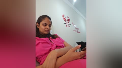 Media: Video of a young woman with medium skin tone, wearing a pink shirt and holding a black phone, sitting in a white room with red heart decals.