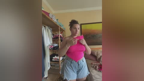 Media: Video of a young woman in a pink, ribbed crop top and frayed denim shorts, standing in a cluttered bedroom with a beige sofa, a colorful sunset painting, and a wooden closet filled with clothes.