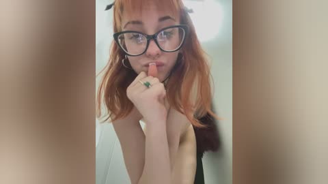 Media: Video of a young woman with fair skin, shoulder-length red hair, and large black-rimmed glasses, topless, biting her finger, standing in a dimly lit room.