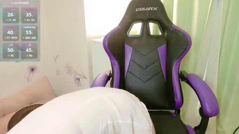 Media: Video of a purple and black GLORIOUS gaming chair in a bright room with a white bed and a green curtain, a wall clock showing 23:45, and a purple wall with flowers.