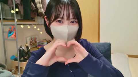 Media: Video of an Asian woman with short black hair, wearing a blue long-sleeve shirt and a white face mask, forming a heart with her hands in a cozy, cluttered room with a desk, shelves, and hanging jackets.