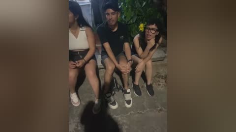 Media: Video of three young adults sitting on a concrete floor, wearing casual clothes. Two are in black T-shirts, one has glasses, and they are surrounded by lush greenery.