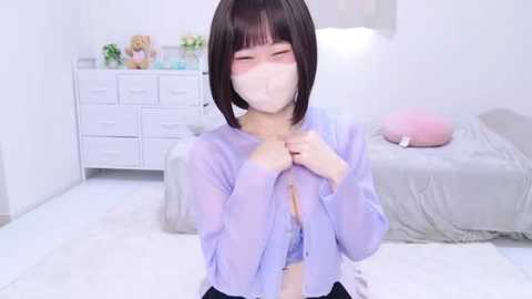 Media: Video of a young Asian woman with a bob haircut, wearing a light purple sheer blouse, blushing, and covering her chest, in a minimalist, white bedroom.