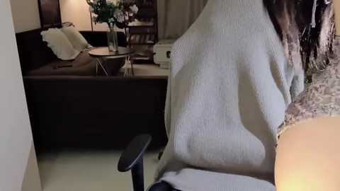 Media: Video of a cozy living room with a beige sweater-wearing woman in a chair, a dark couch with pillows, and a wooden coffee table holding a vase of flowers.