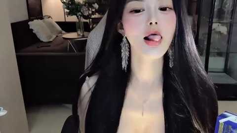 Media: Video of an East Asian woman with long black hair, wearing a low-cut top, blushing cheeks, and sticking out her tongue. Background shows a modern living room with a dark sofa, flowers, and a glass door.