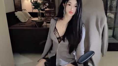 Media: Video of an Asian woman with long black hair, wearing a gray cardigan over a black lace bra, sitting on a beige floor, looking into a mirror, in a modern, dimly-lit living room.